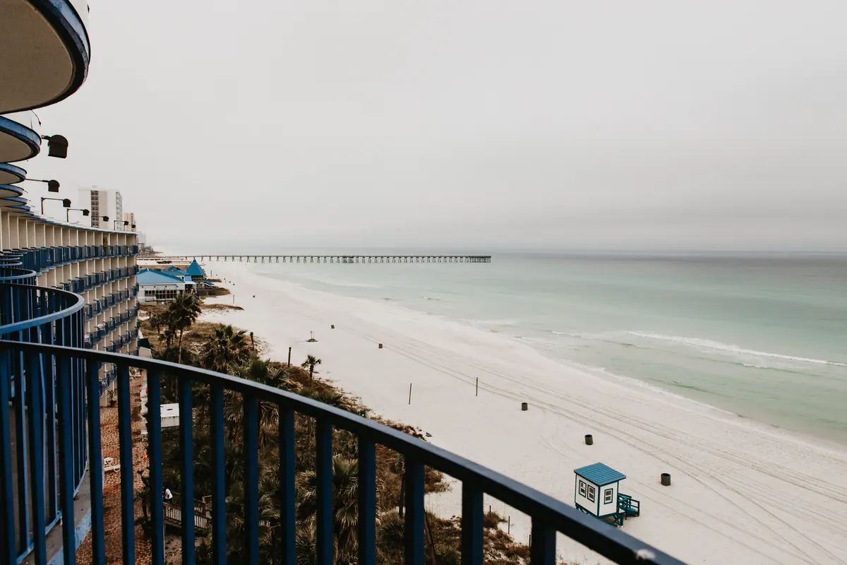 Curved blue balcony railing high above a white-sand beach with a lifeguard stand, a long fishing pier and pale green water