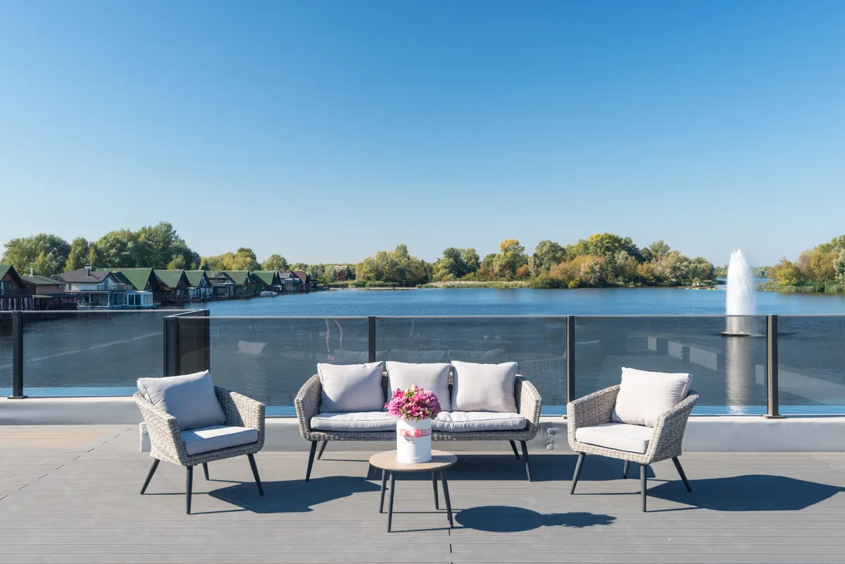 Outdoor lounge chairs and a small table on a terrace with a glass railing overlooking a lake and a fountain