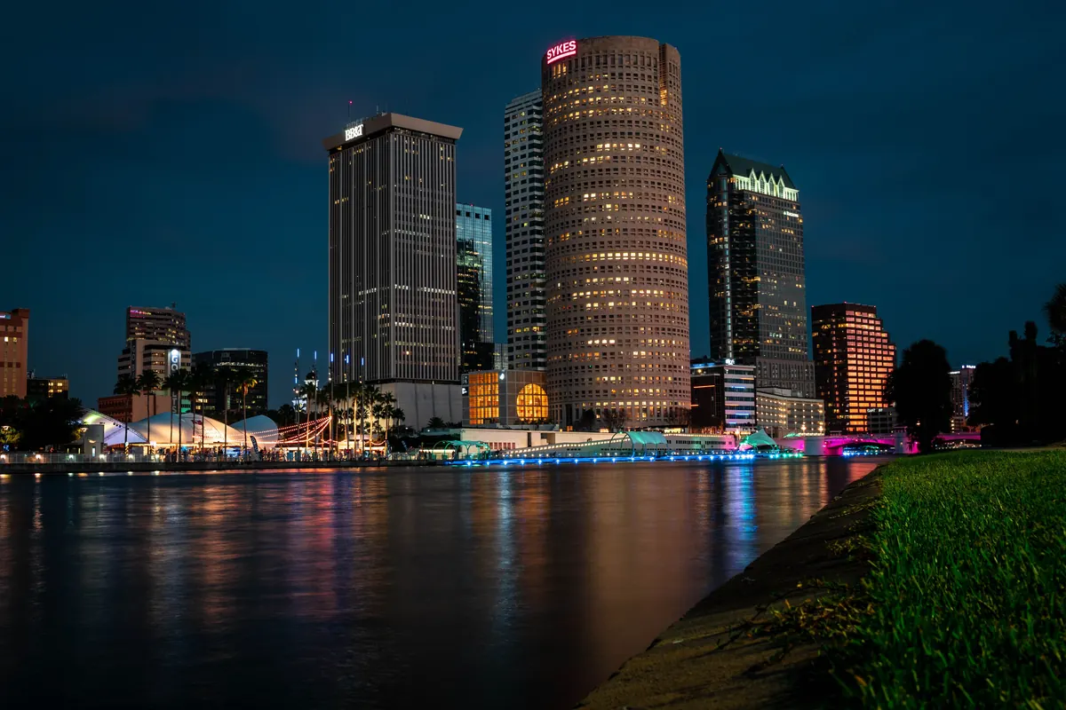 Downtown Tampa towers lit at blue hour across the Hillsborough River, reflections on the water and a grassy bank in the foreground